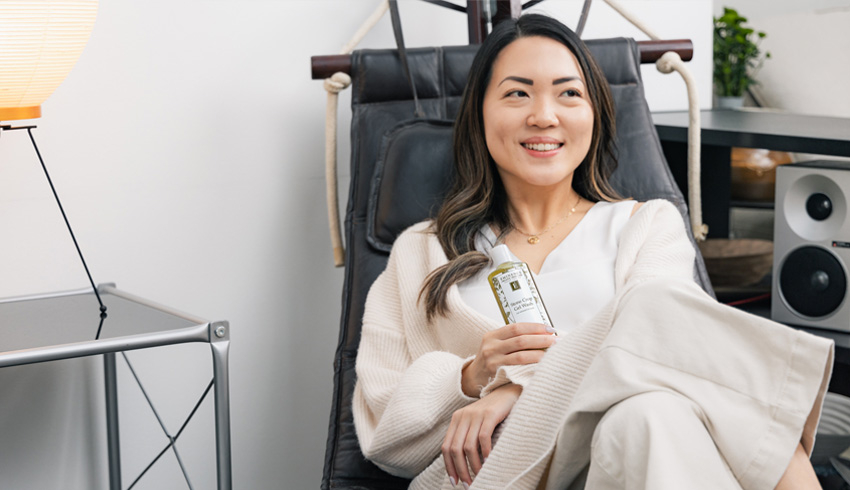 A woman sitting down in a chair and holding an Eminence Organics product in one hand. 
